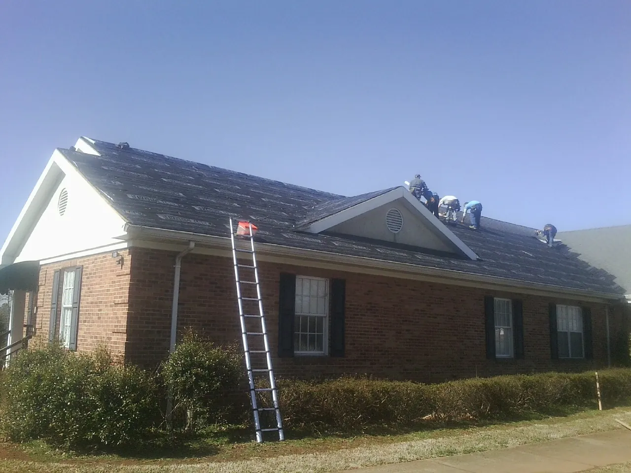 Crew tearing shingles off a brick ranch roof with a ladder set — Emergency Roof Repair work by our crew in Middle Amana